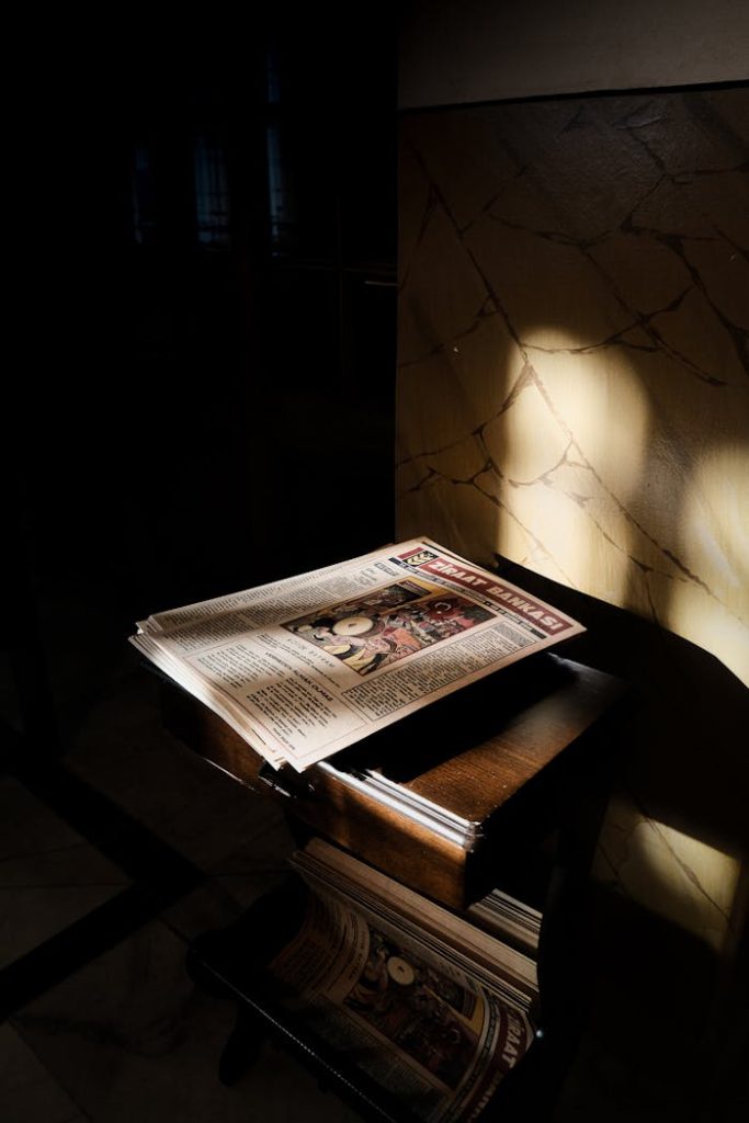 A newspaper bathed in sunlight rests on a wooden table in a dimly lit room.